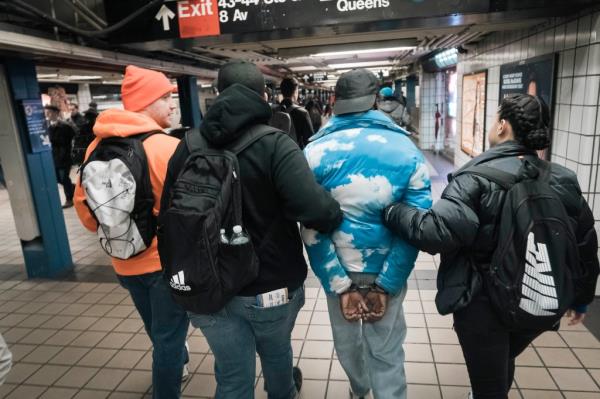 A man in handcuffs seen from behind with police in plain clothes holding him by his arms, walking through a subway concourse