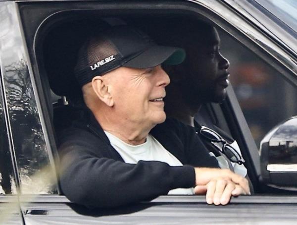 Bruce Willis, wearing a black jacket, white top and black cap, smiles in a car during an outing 